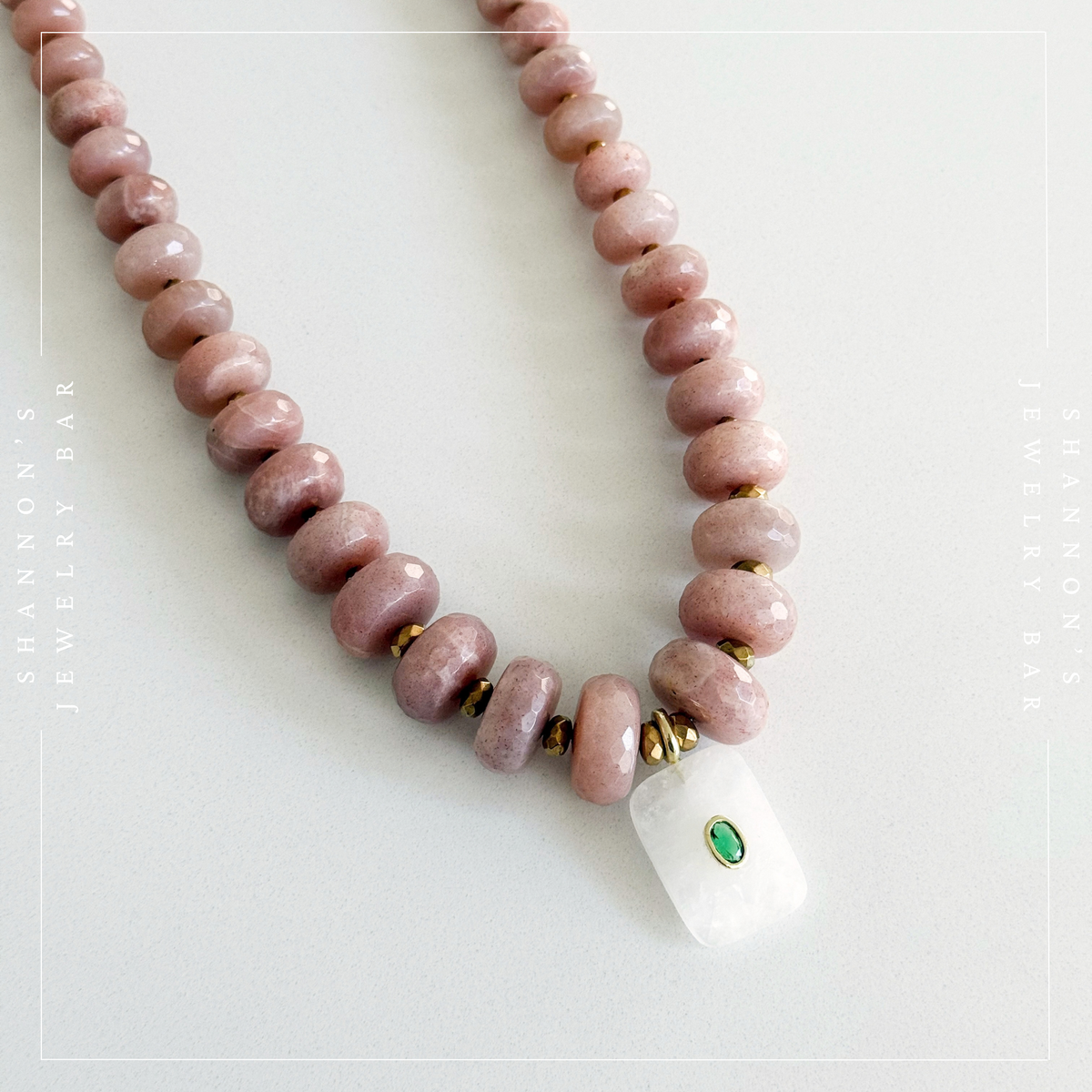 A beaded necklace featuring polished dusty pink stone beads with small gold spacer rings and a white rectangular pendant with a green oval accent, displayed on a light gray background. Peach Moonstone, Faceted Graduated Beads.