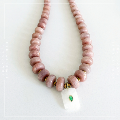 A beaded necklace made of dusty pink stone beads with a white rectangular pendant featuring a small green stone and gold bezel, displayed on a light gray background.  Peach Moonstone, Faceted Graduated Beads.