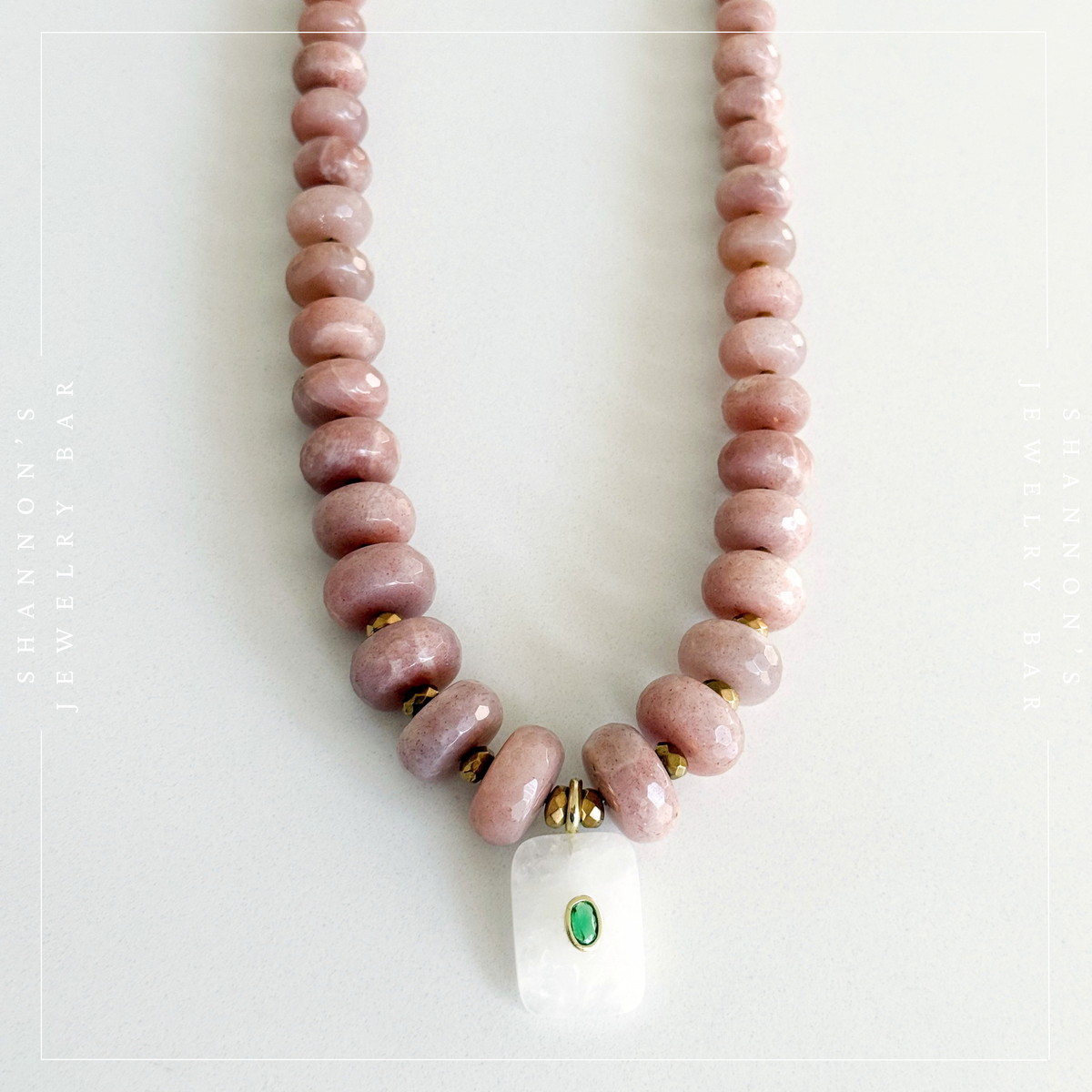 A beaded necklace featuring dusty pink stone beads with small gold spacer accents and a white rectangular pendant with a green oval detail, displayed on a light off-white background.  Peach Moonstone, Faceted Graduated Beads.