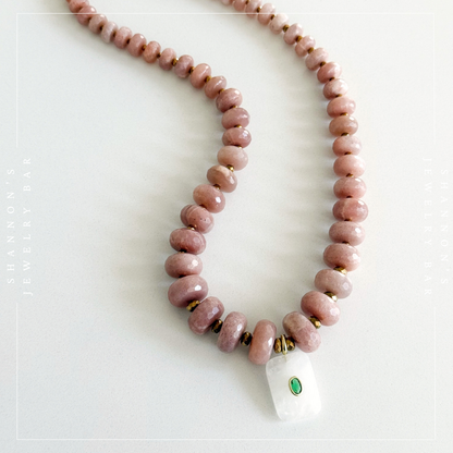 A beaded necklace made of polished dusty pink stone beads with small gold spacer rings and a white rectangular pendant featuring a green oval inlay, displayed on a light background.  Peach Moonstone, Faceted Graduated Beads.