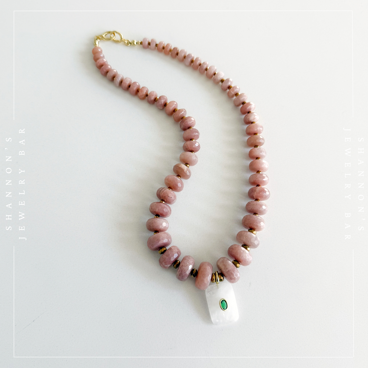 A beaded necklace with round dusty pink beads, small gold spacer beads, and a white rectangular pendant featuring a green inlay, displayed on a light gray background.  Peach Moonstone, Faceted Graduated Beads.