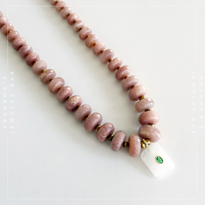 A beaded necklace with dusty pink stone beads, small gold spacer beads, and a white rectangular pendant featuring a green inlay, displayed on a light gray background.  Peach Moonstone, Faceted Graduated Beads.
