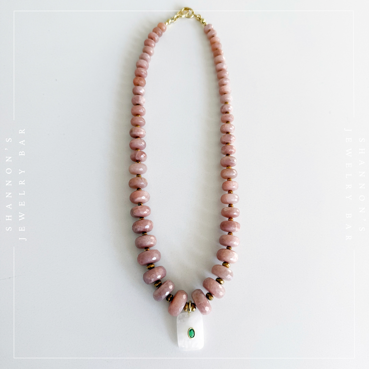 A beaded necklace made of pinkish stone beads with small gold spacer rings and a white rectangular pendant featuring a green stone, displayed on a light gray background.  Peach Moonstone, Faceted Graduated Beads.