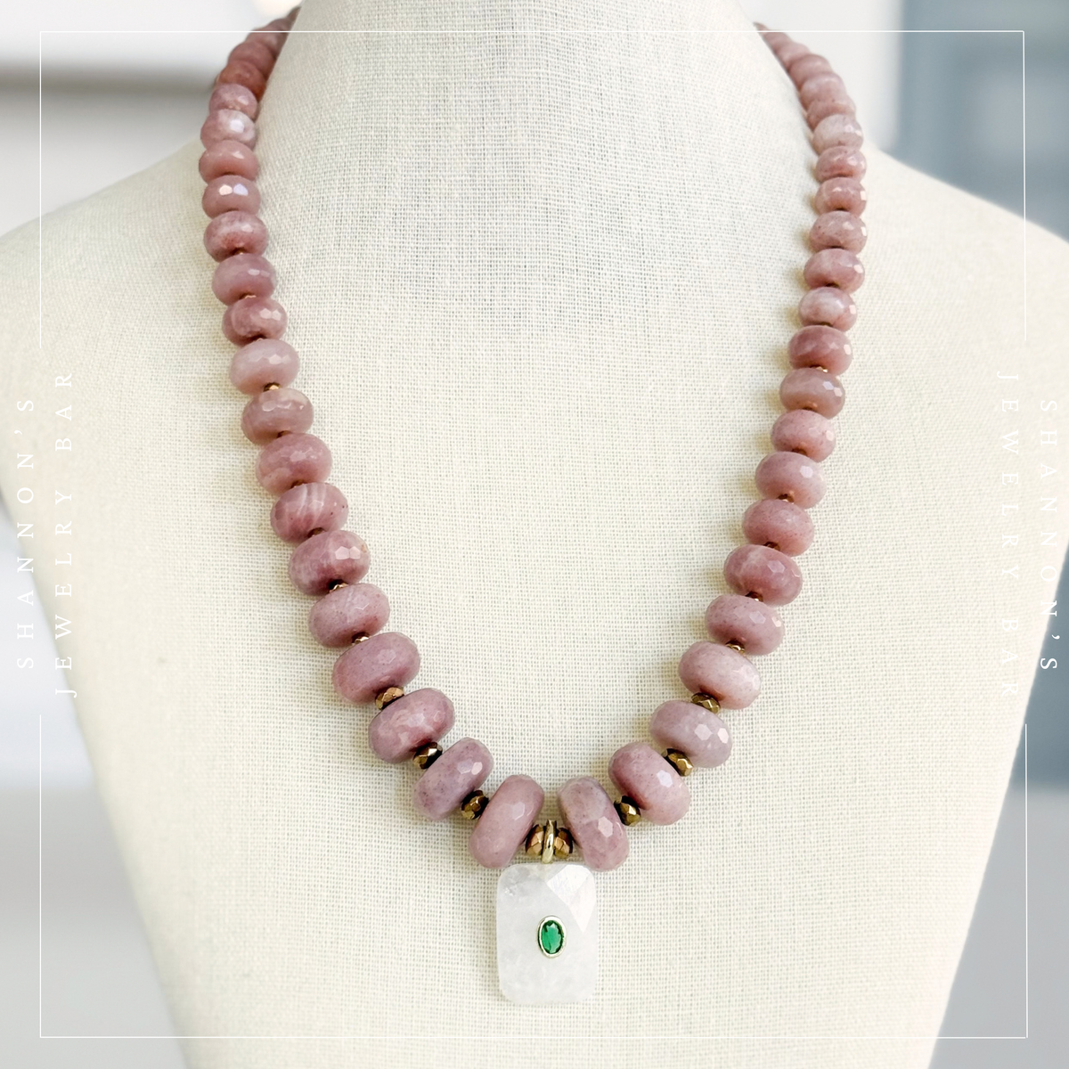 A beaded necklace displayed on a cream fabric jewelry bust. The necklace features dusty pink faceted beads, small gold spacer rings, and a white rectangular pendant with a small green stone accent. Peach Moonstone, Faceted Graduated Beads.
