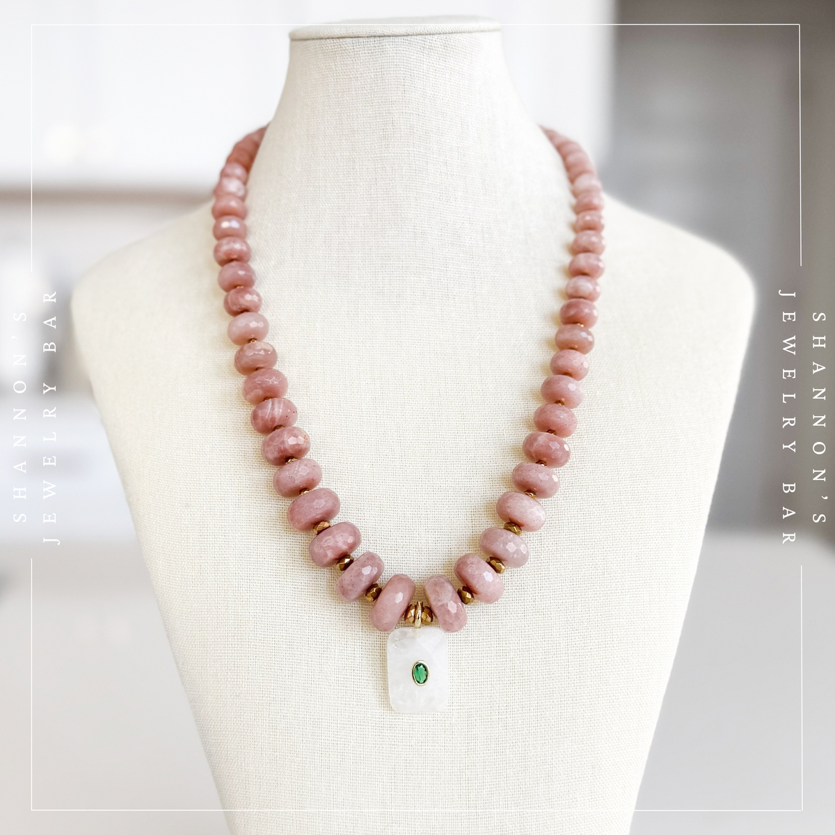 A pink stone beaded necklace with small gold spacer beads and a white pendant featuring a green stone, displayed on a cream fabric jewelry mannequin bust. Peach Moonstone, Faceted Graduated Beads.