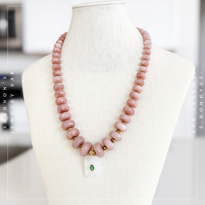 A pink stone beaded necklace displayed on a cream fabric jewelry mannequin bust. The necklace features irregular dusty pink beads with small gold spacer rings and a white pendant containing a small green stone. Peach Moonstone, Faceted Graduated Beads.