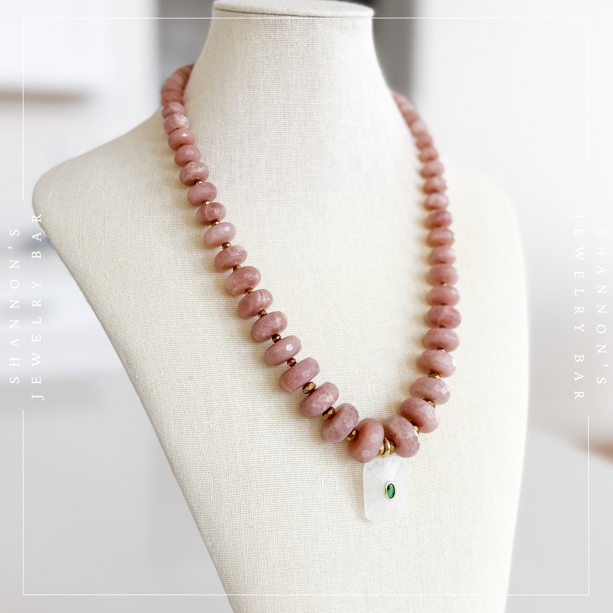 A pink stone beaded necklace with small gold spacer beads and a white rectangular pendant featuring a green stone, displayed on a cream fabric jewelry bust.  Peach Moonstone, Faceted Graduated Beads.