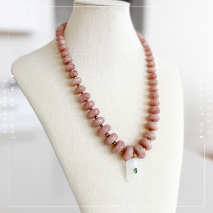 A pink stone beaded necklace with small gold spacer beads and a white rectangular pendant featuring a green stone, displayed on a cream fabric jewelry bust.  Peach Moonstone, Faceted Graduated Beads.