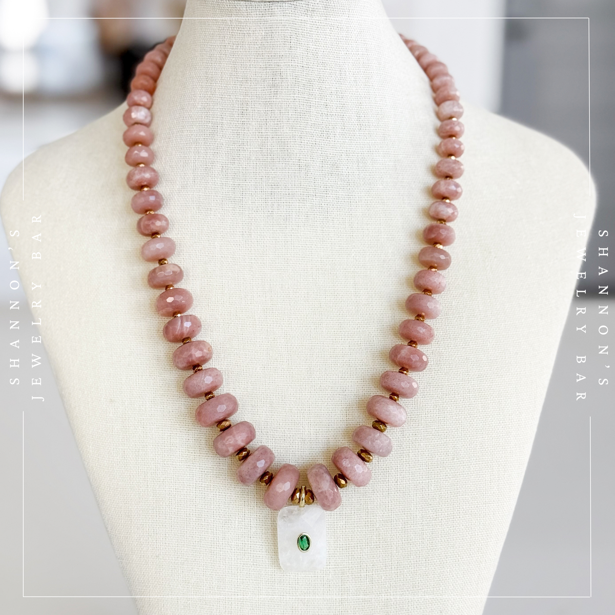 A pink stone bead necklace displayed on a cream fabric jewelry bust. The necklace features round dusty pink beads, small gold spacer beads, a white rectangular pendant with a green stone, and gold metal accents.  Peach Moonstone, Faceted Graduated Beads.