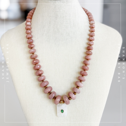 A pink stone bead necklace displayed on a cream fabric jewelry bust. The necklace features round dusty pink beads, small gold spacer beads, a white rectangular pendant with a green stone, and gold metal accents.  Peach Moonstone, Faceted Graduated Beads.