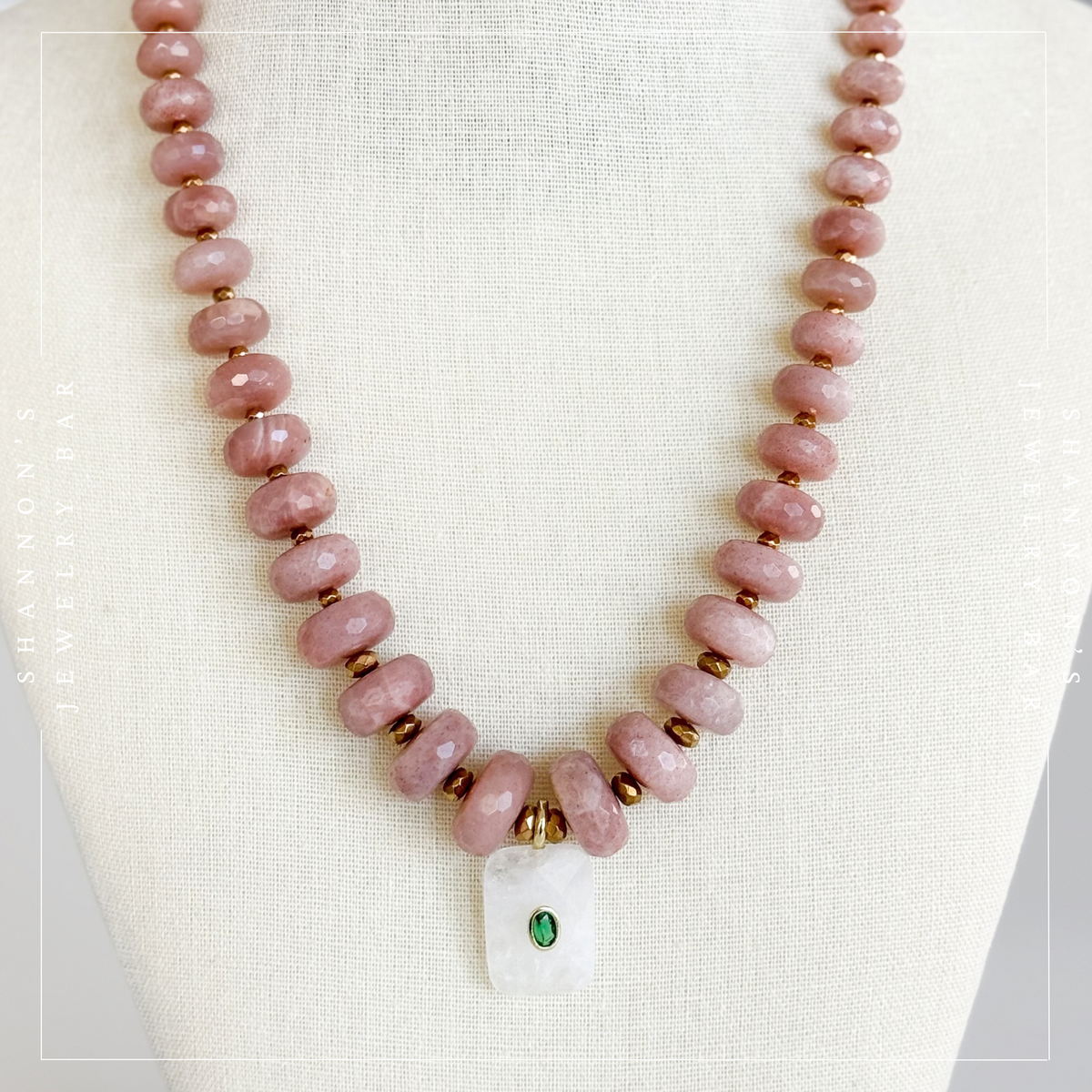 A beaded necklace with chunky dusty pink stone beads, small gold spacer beads, and a white rectangular pendant featuring a green oval stone, displayed on a cream fabric jewelry bust.  Peach Moonstone, Faceted Graduated Beads.