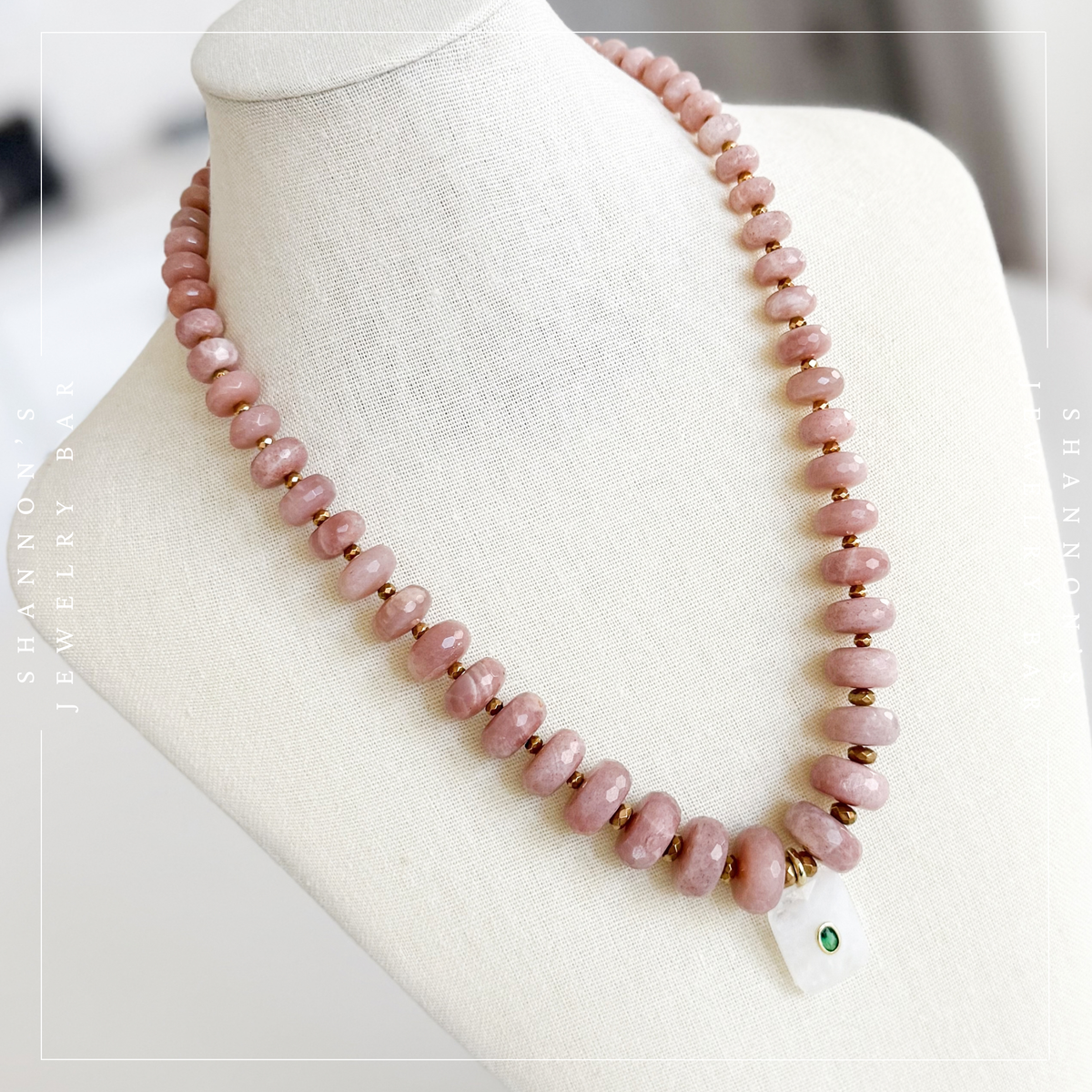 A pink stone bead necklace displayed on a cream fabric jewelry bust. The necklace features small round pink beads separated by tiny gold spacer beads, with a white rectangular pendant at the front secured by gold wire and a small green stone accent.  Peach Moonstone, Faceted Graduated Beads.